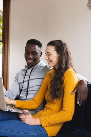 Happy diverse couple sitting on sofa using laptop at home. Lifestyle, togetherness, relationship, communication and dometic life, unaltered.の写真素材