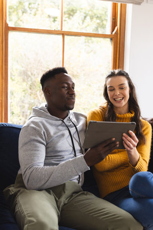 Happy diverse couple sitting on sofa using tablet at home. Lifestyle, togetherness, relationship,...