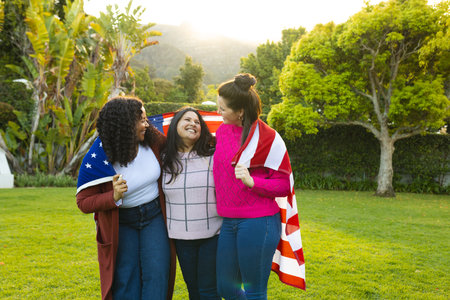 Three happy diverse female friends with flag on backs in sunny garden. Celebration, friendship,...