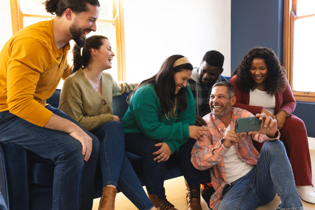 Happy diverse male and female friends relaxing at home together talking and looking at smartphone. Communication, lifestyle, friendship, relaxation, free time and domestic life, unalteredの写真素材