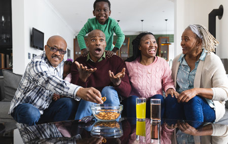 Excited african american parents, son and grandparents watching sport on tv, slow motion. Sport, entertainment, home, family, togetherness, domestic life and lifestyle, unaltered.の写真素材