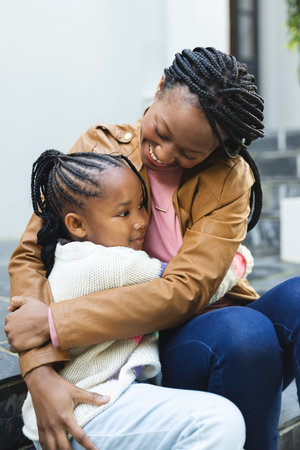 Happy african american mother and daughter embracing on steps in front of house. Home, family, domestic life and lifestyle, unaltered.の写真素材