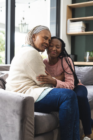 Happy african american senior mother and adult daughter sitting on couch and embracing. Home, family, togetherness, domestic life and lifestyle, unaltered.の写真素材