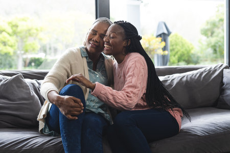 Happy african american senior mother and adult daughter sitting on couch,laughing and embracing. Home, family, togetherness, domestic life and lifestyle, unaltered.の写真素材