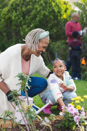 Happy african american grandmother and granddaughter watering plants in sunny garden. Family,...