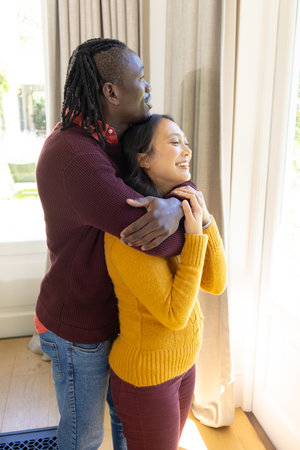 Happy diverse couple standing by window embracing and smiling in sunny living room, copy space. Love, free time, togetherness, relationship, relaxation, domestic life and lifestyle, unaltered.の写真素材