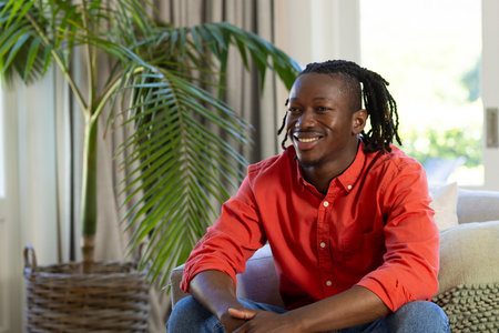 Happy african american man with dreadlocks sitting on couch smiling in sunny living room. Free time,...
