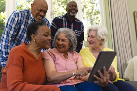 Happy diverse group of senior friends using tablet and laughing in sunny living room. Retirement, friendship, domestic life and senior lifestyle, communication, unaltered.の写真素材