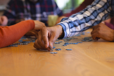 Hands of diverse group of senior friends playing with jigsaw puzzles in sunny dining room at home. Retirement, friendship, wellbeing, activities, togetherness and senior lifestyle, unaltered.の写真素材