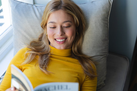 Happy caucasian woman lying on couch, reading book and smiling in sunny room at home. Relaxation, wellbeing, free time and domestic life, unaltered.の写真素材