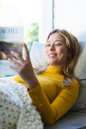 Happy caucasian woman lying on couch, reading book and smiling in sunny room at home. Relaxation, wellbeing, free time and domestic life, unaltered.の写真素材