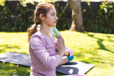 Focused biracial woman practicing yoga meditation sitting with friend in sunny garden, copy space. Summer, yoga, fitness and healthy lifestyle, unaltered.の写真素材