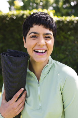 Portrait of happy biracial woman holding yoga mat smiling in sunny garden. Summer, nature, fitness and healthy lifestyle, unaltered.の写真素材
