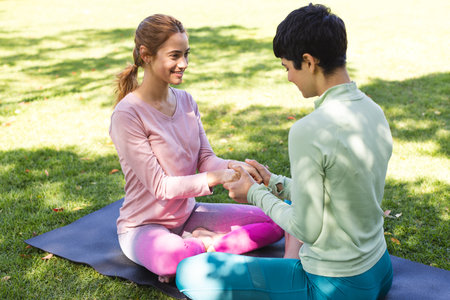 Happy biracial lesbian couple practicing yoga sitting holding hands and smiling in sunny garden....