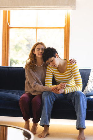 Sad biracial lesbian couple embracing and comforting, on couch in sunny living room, copy space....
