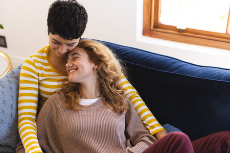 Happy biracial lesbian couple relaxing on couch embracing together at home. Gay, love, relationship, togetherness, domestic life and lifestyle, unaltered.の写真素材