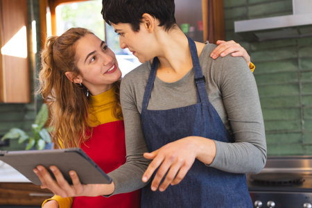 Happy biracial lesbian couple preparing food, using tablet and embracing in kitchen. Communication, cooking, food, gay, relationship, togetherness, domestic life and healthy lifestyle, unaltered.の写真素材