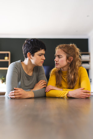 Angry biracial lesbian couple at dining table having video call, looking at each other, copy space. Communication, technology, gay, relationship, togetherness, domestic life and lifestyle, unaltered.の写真素材