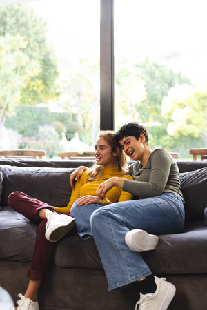 Happy biracial lesbian couple sitting on couch laughing in sunny living room, copy space. Relaxation, gay, relationship, togetherness, domestic life and lifestyle, unaltered.の写真素材