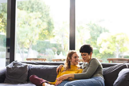 Happy biracial lesbian couple sitting on couch talking in sunny living room, copy space. Relaxation, gay, relationship, togetherness, domestic life and lifestyle, unaltered.の写真素材
