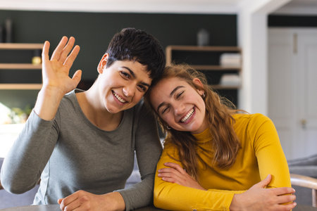Happy biracial lesbian couple sitting at home having video call, waving. Communication, technology,...