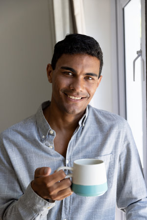 Portrait of happy biracial man holding mug next to window at home. Lifestyle and domestic life, unaltered.の写真素材