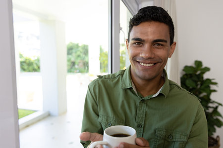 Portrait of happy biracial man holding mug standing next to window at home. Lifestyle and domestic life, unaltered.の写真素材