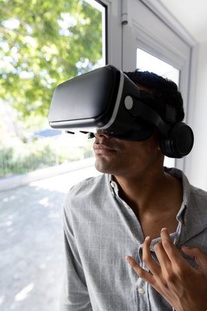 Biracial man using vr headset next to window at home. Technology, communication, lifestyle and...