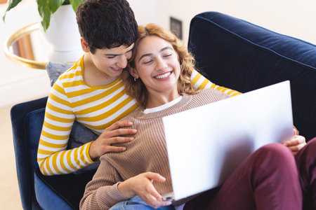 High angle of happy biracial lesbian couple relaxing on couch using laptop together. Communication, free time, relaxation, relationship, togetherness, domestic life and lifestyle, unaltered.の写真素材