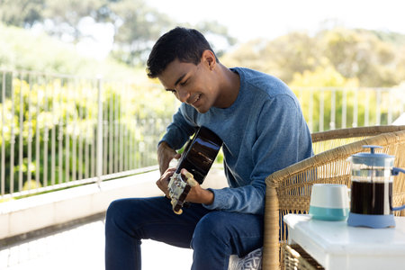 Happy biracial man sitting on balcony playing guitar at home. Hobbies, music, lifestyle and domestic life, unaltered.の写真素材