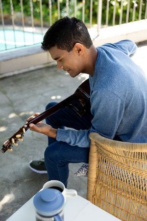 Happy biracial man sitting on balcony playing guitar at home. Hobbies, music, lifestyle and domestic...