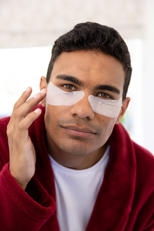 Happy biracial man applying uder-eye patches in bathroom at home. Self care, lifestyle and domestic...