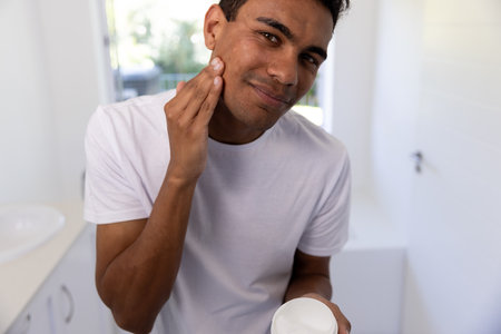 Happy biracial man applying face cream in bathroom at home. Self care, lifestyle and domestic life, unaltered.の写真素材