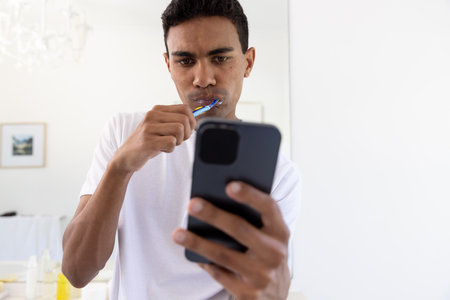 Biracial man brushing teeth and using smartphone in bathroom at home. Self care, hygiene, communication, lifestyle and domestic life, unaltered.の写真素材