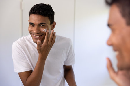 Happy biracial man applying face cream looking in mirror in bathroom at home. Self care, lifestyle and domestic life, unaltered.の写真素材