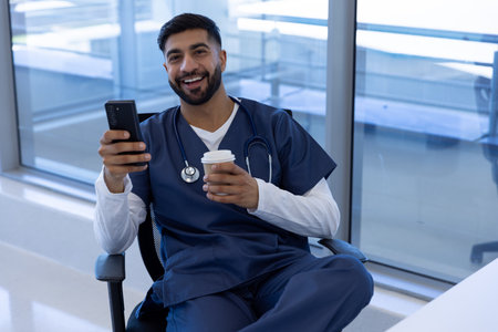 Portrait of happy biracial male doctor using smartphone and holding cup of coffee in hospital office. Medicine, healthcare, communication and medical services, unaltered.の写真素材