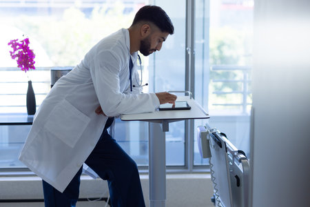Biracial male doctor using tablet in sunny hospital room. Medicine, healthcare, communication and...