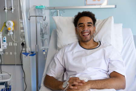 Portrait of happy biracial male patient in bed in sunny hospital room. Medicine, healthcare and medical services, unaltered.の写真素材