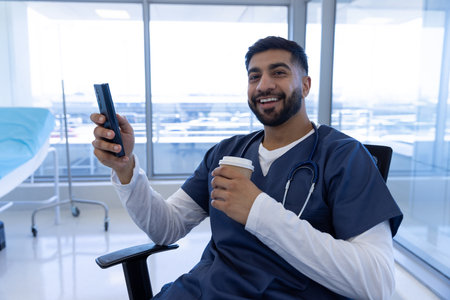 Portrait of happy biracial male doctor using smartphone and holding cup of coffee in hospital...