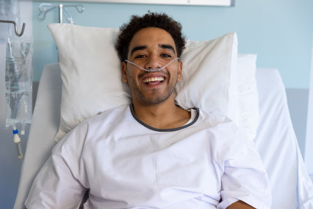 Portrait of happy biracial male patient in bed in sunny hospital room. Medicine, healthcare and medical services, unaltered.の写真素材