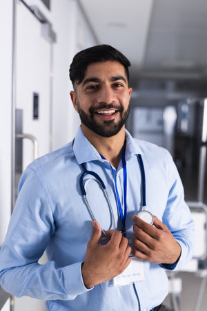 Portrait of happy biracial male doctor with stethoscope in hospital corridor. Medicine, healthcare and medical services, unaltered.の写真素材