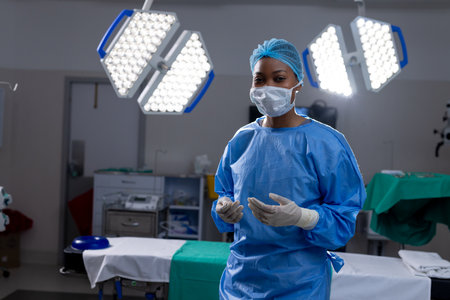 Portrait of african american female doctor with face mask and gloves in hospital operating room....