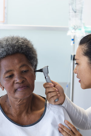 Diverse female doctor testing ear of senior female patient in hospital room. Medicine, healthcare and medical services, unaltered.の写真素材