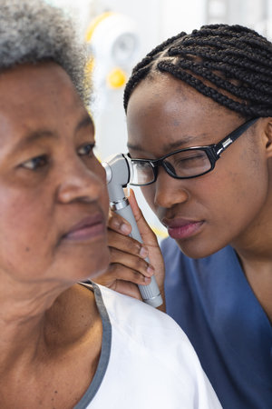 African american female doctor testing ear of senior female patient in hospital room. Medicine, healthcare and medical services, unaltered.の写真素材