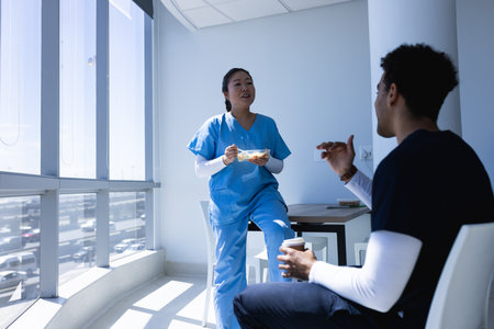 Diverse male and female doctors talking, drinking coffee and eating lunch in sunny hospital....