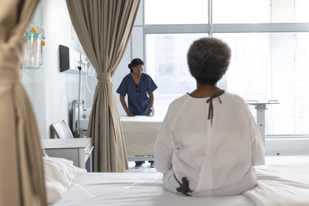 Back of african american senior female patient sitting on bed in sunny hospital room. Medicine,...