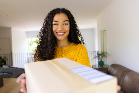 Happy biracial woman in yellow sweater holding package and using smartphone at home. Lifestyle, free time, communication and domestic life, unaltered.の写真素材