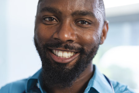 Portrait of happy african american male doctor wearing blue shirt in hospital room. Medicine,...