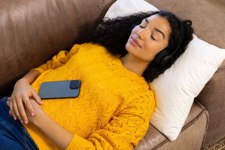 Biracial woman with headphones and smartphone lying on sofa at home. Lifestyle, free time,...