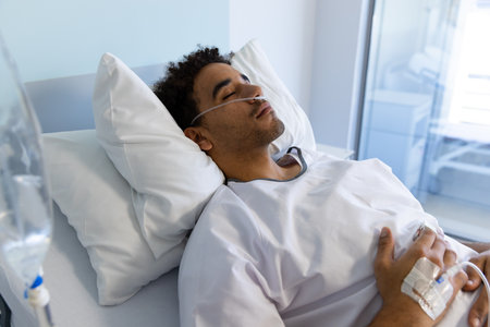 Biracial male patient sleeping in bed with iv drip in sunny hospital room. Medicine, healthcare and...